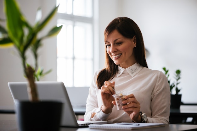 Cheerful businesswoman sitting in office with a laptop having dessert.