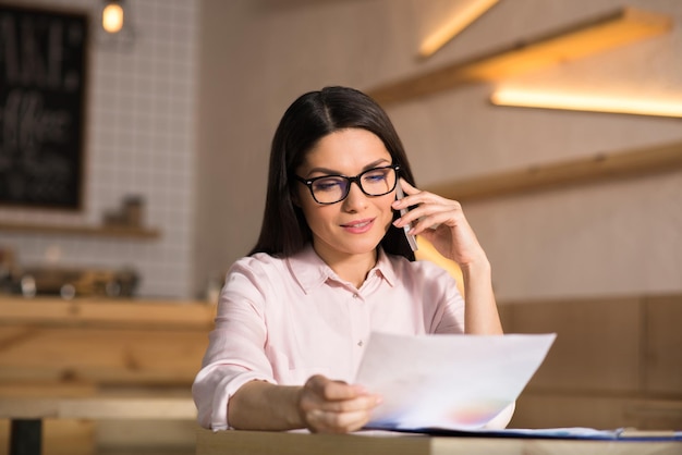 Cheerful businesswoman wearing eyeglasses talking on smartphone and holding document in cafe