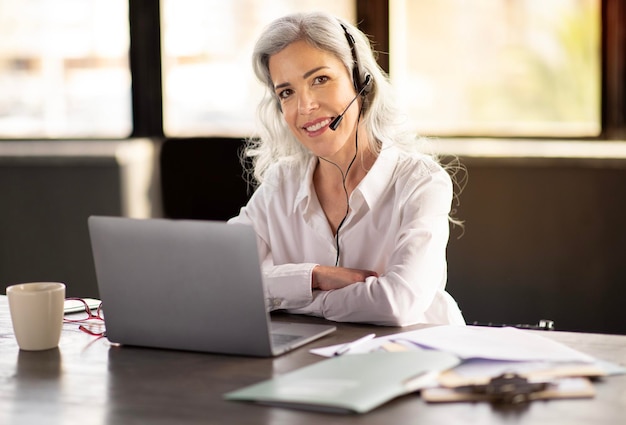 Веселая деловая женщина в наушниках общается онлайн на ноутбуке в офисе
