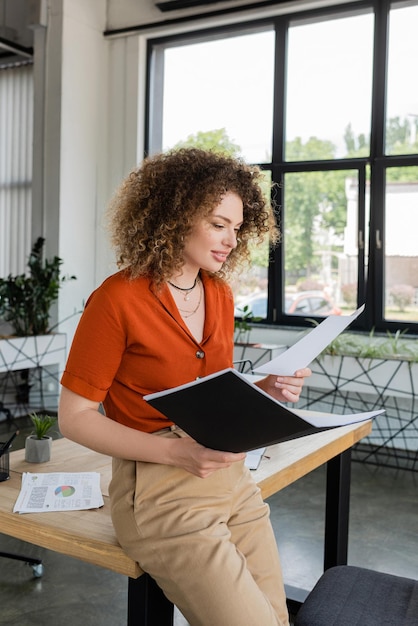 cheerful businesswoman with curly hair looking at document and holding folder in office