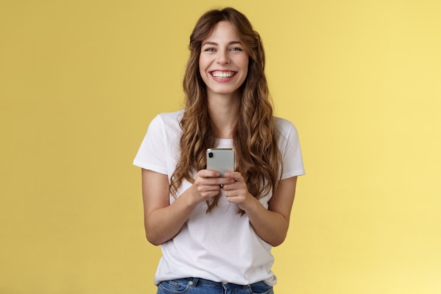 Cheerful carefree happy lively girl hold smartphone using awesome funny app playing cool game relaxing waiting friend park browsing social media using mobile phone look camera upbeat smiling