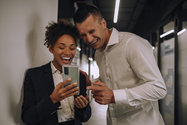 Cheerful case Laughing young adult elegant mulatta woman and caucasian man looking at smartphone screen standing in office corridor