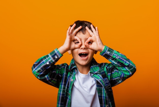 Cheerful Caucasian boy holding hands near face in the form of a mask, isolated on yellow background.