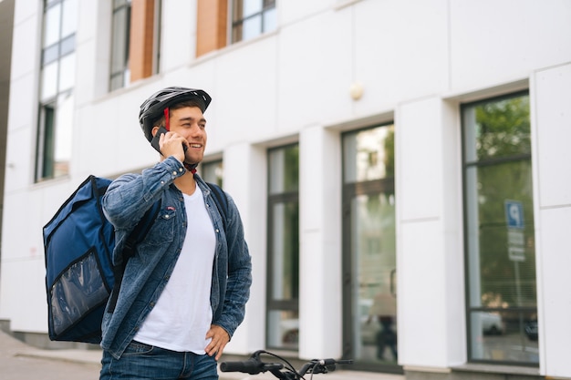 Corriere caucasico allegro maschio con zaino termico che parla al telefono cellulare con il cliente, cercando l'indirizzo per consegnare l'ordine online. fattorino in casco protettivo utilizzando smartphone all'aperto.