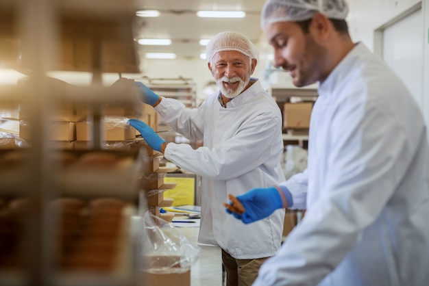 食品工場に立ちながら、白い無菌の制服を着た陽気な白人の従業員が箱にクッキーを詰めました。