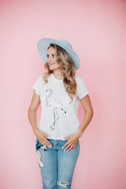 Cheerful caucasian female with long wavy fair hair in white t-shite, blue jeans, big blue hat smiles