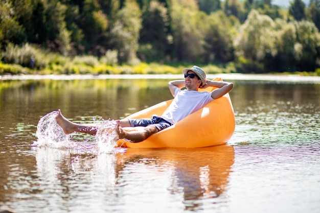 Cheerful caucasian good looking happy guy in sunglasses sitting on orange inflatable mattress with h