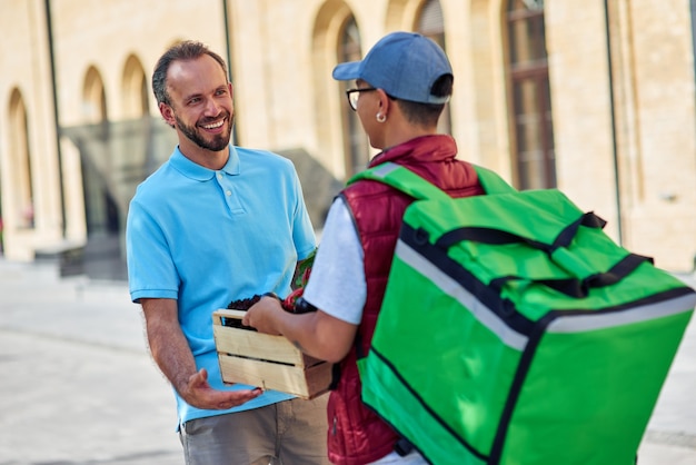 Photo cheerful caucasian man customer receiving wooden box with fresh grocery from male courier with