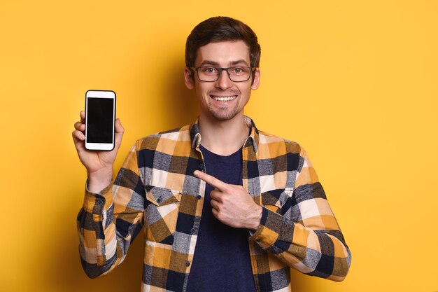 Photo cheerful caucasian man in glasses pointing with finger at blank smartphone display in his hand, smiling, looking at the camera, chatting, advice, advertising. isolated on yellow