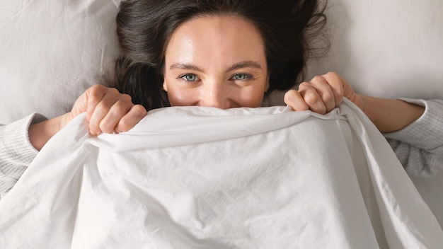 Cheerful caucasian millennial female lies on comfortable bed enjoys softness weekend vacation and free time
