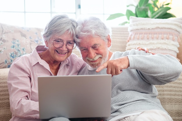 Cheerful caucasian senior couple sitting on the floor at home using laptop modern retired elderly people surfing the net with computer