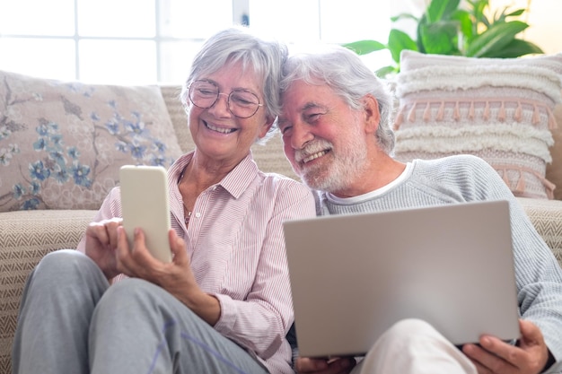 노트북과 스마트 폰을 사용하여 집 바닥에 앉아 있는 쾌활한 백인 노인 부부는 컴퓨터로 인터넷을 서핑하는 현대 은퇴한 노인들