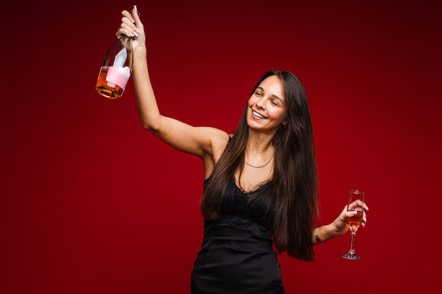 Cheerful caucasian woman with attractive appearance with a bottle of champagne and glass, picture isolated on red wall