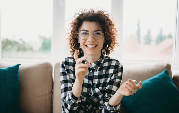 Cheerful caucasian woman with curly hair and eyeglasses smiling at camera while sitting near the window on the sofa