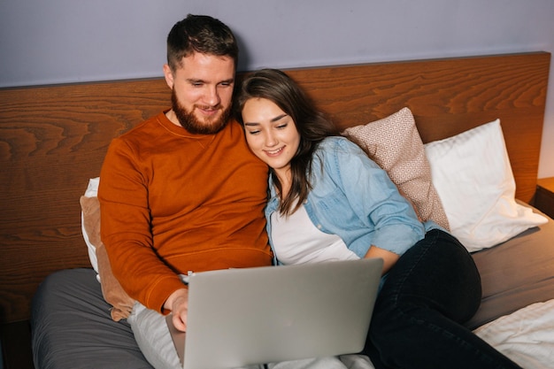 Cheerful Caucasian young couple watching video online on laptop lying in bed at night cuddling and watching movie on computer