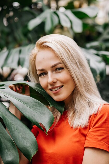 Cheerful charming caucasian blonde woman wearing red t shirt enjoying time in forest, smiling with white teeth standing among tropical leaves. Nature and people concept.