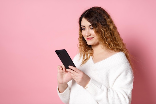 A cheerful charming young woman standing isolated on a pink background with a smartphone in hands