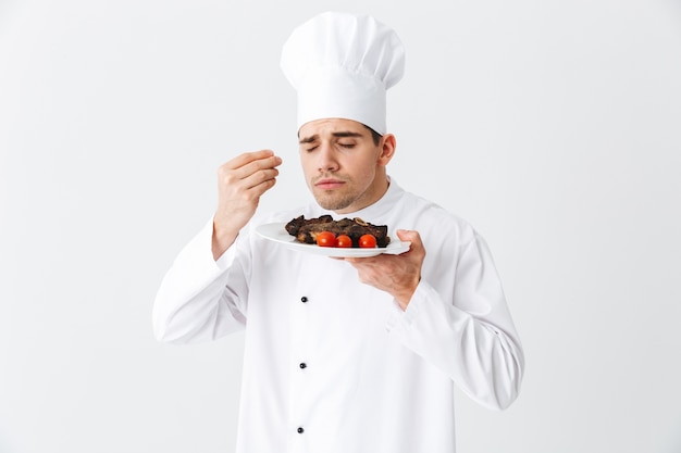 Cheerful chef cook wearing uniform peppers cooked beef steak meal on a plate isolated over white wall