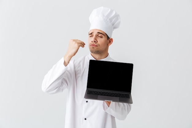 Cheerful chef cook wearing uniform standing over white wall, holding bank screen laptop computer