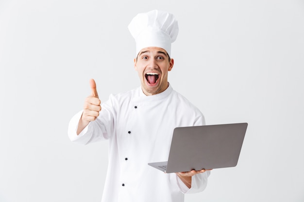 Allegro chef cuoco indossa uniforme in piedi sul muro bianco, tenendo il computer portatile