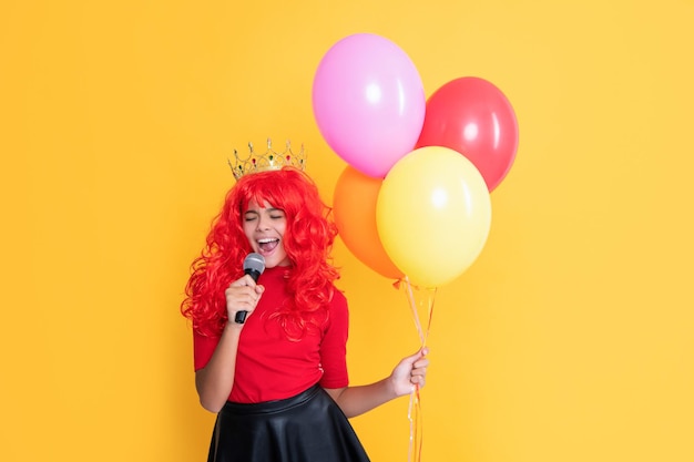 Cheerful child in crown with microphone and party balloon on yellow background