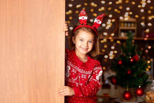 Una bambina allegra guarda fuori da dietro la porta in una cucina buia con un albero di natale per capodanno o natale, sorridendo e ridendo con un cappello di babbo natale, spazio per il testo