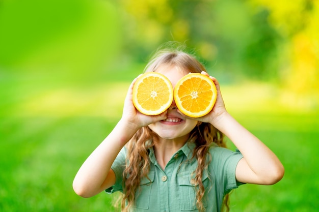 A cheerful child girl in the summer on the lawn covered her face with oranges on the green grass has fun and rejoices space for text