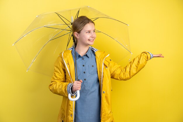 Ragazza allegra del bambino con l'ombrello e il cappotto di pioggia giallo sulla parete gialla