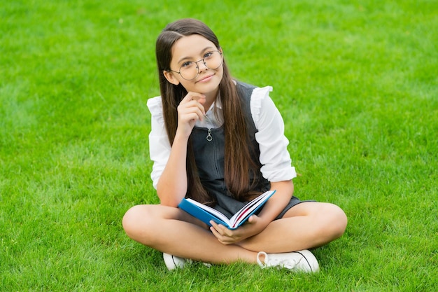 Cheerful child in glasses reading book sitting on green grass