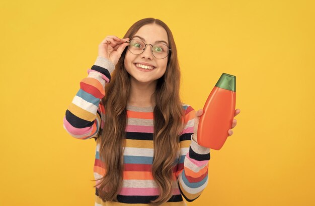 Cheerful child long hair in glasses presenting shampoo bottle advertisement