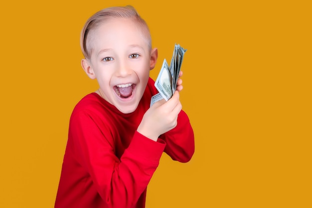 Photo a cheerful child in red holds a bundle of dollar bills in front of him and makes funny faces