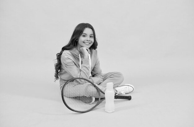 Cheerful child sit in sportswear with badminton racquet and water bottle on yellow background relax