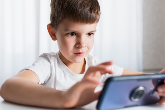 A cheerful child in a white T-shirt plays games on the phone at home. Happy boy looks at his smartphone.