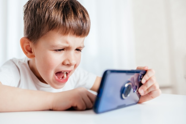 Веселый ребенок в белой футболке играет дома по телефону. Счастливый мальчик смотрит на свой смартфон.