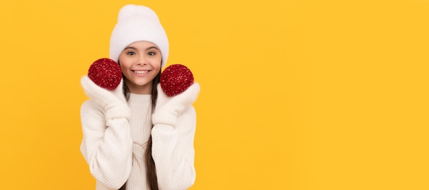 Photo cheerful child in winter hat and gloves hold decorative xmas balls on yellow background xmas banner of christmas child girl studio kid winter portrait with copy space