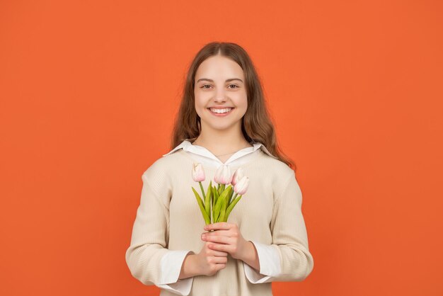 チューリップと元気な子。子供は春休みに花を持っています。オレンジ色の背景に花束を持つ十代の少女。子供の頃の幸せ。女性の日のための花のプレゼント。 3月の日。