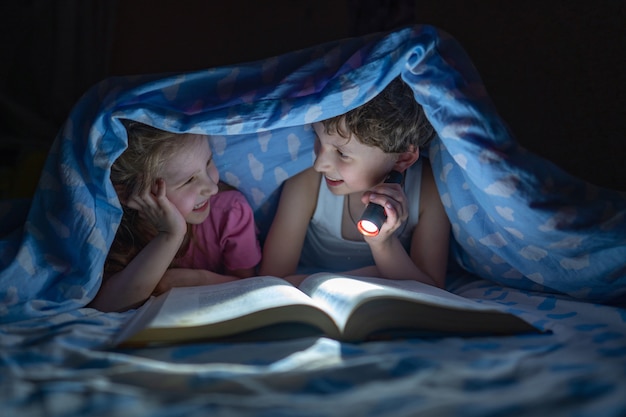 Cheerful children, brother and sister lie under the covers and read a book