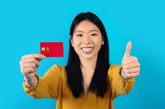 Cheerful chinese young woman showing bank card on blue