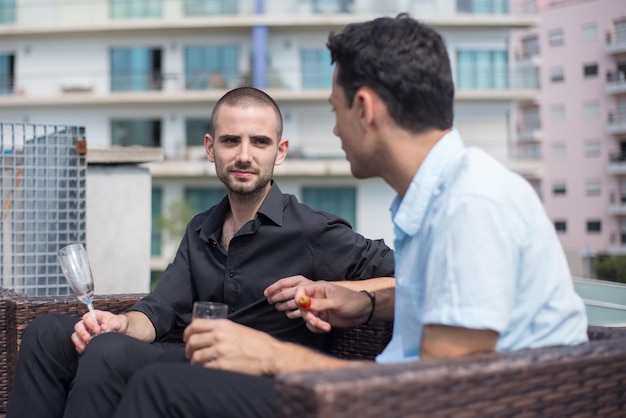 Cheerful colleagues on terrace roof party. Men in formal clothes sitting on rattan sofas with champagne, eating appetizers. Teambuilding, party concept