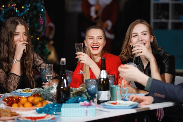 Cheerful company of friends celebrates the New Year at a table near the Christmas tree with garlands. Women and men laugh, rejoice with champagne glasses.