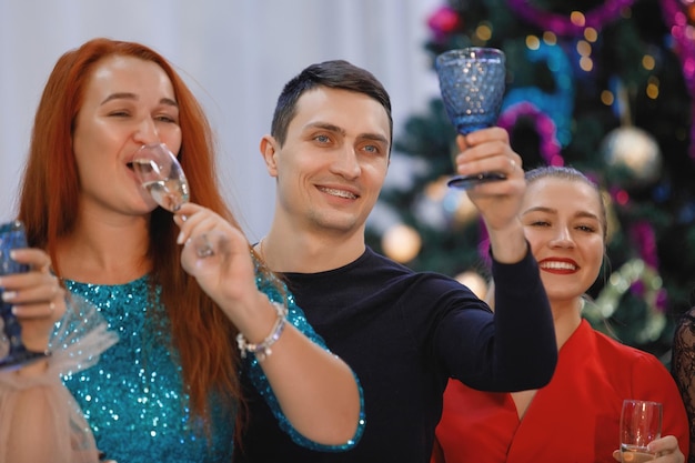 Фото Веселая компания друзей встречает новый год за столом возле елки с гирляндами. женщины и мужчины смеются, радуются бокалам с шампанским.