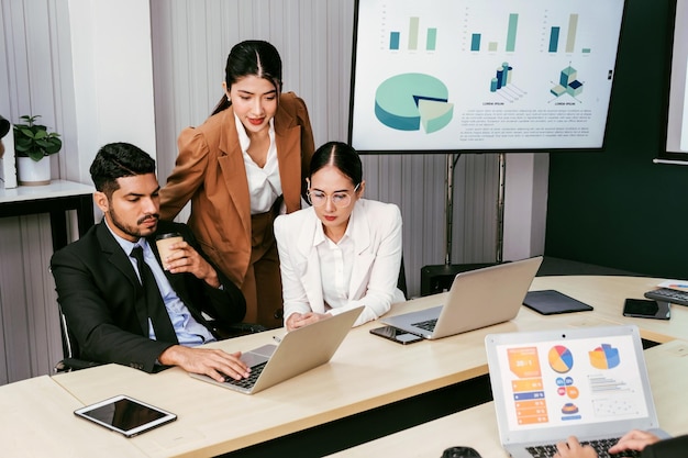 A cheerful and confident Asian businesswoman stands in the back of her office colleagues presenting bar charts data from a monitor and projector screen Asian businesswoman leader role at the meeting