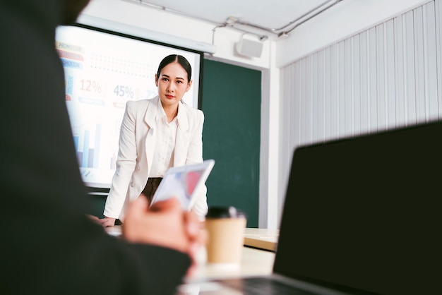 A cheerful and confident Asian businesswoman stands present bar charts data from projector screen to her office colleagues Asian business women leader role at the meeting