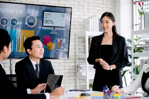 A cheerful and confident Asian businesswoman stands present bar charts data from a whiteboard to her office colleagues Asian business women leader role at the meeting