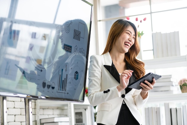 A cheerful and confident Asian businesswoman stands and uses a tablet to present a bar chart of data from a monitor to her office colleagues Focus on Asian business women leader role at the meeting