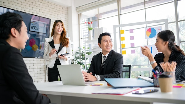 A cheerful and confident Asian businesswoman stands and uses a tablet to present a bar chart of data from a monitor to her office colleagues Focus on Asian business women leader role at the meeting