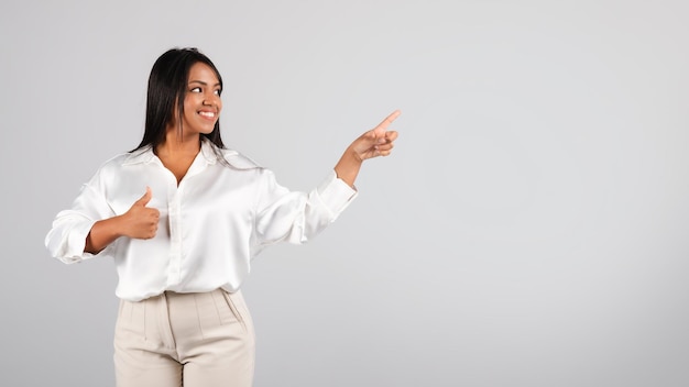 Cheerful confident millennial black businesswoman in white blouse point fingers at empty space