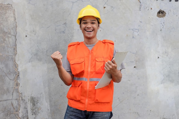 Cheerful contractor man in helmet holds clipboard and gesturing raise arm as achievement sign