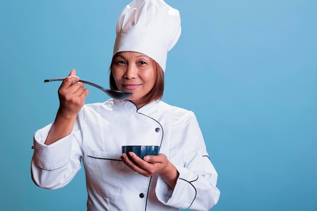 Cheerful cook with restaurant uniform holding bowl with culinary meal while tasting food before start eating, cooking healthy recipe. Professional asian chef preparing dinner dish, gastronomy industry