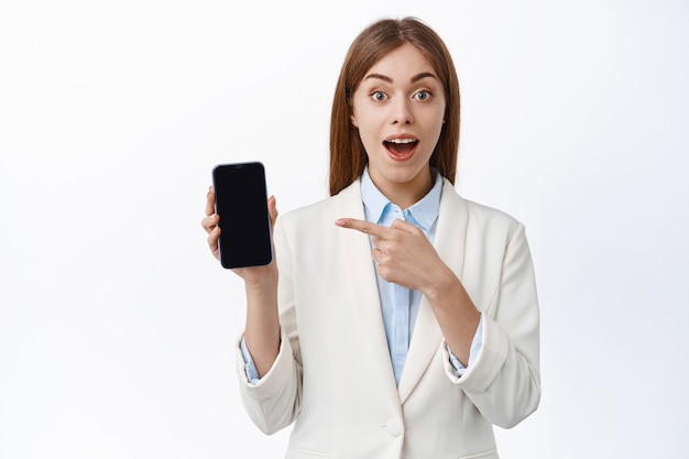 Cheerful corporate woman in business suit, points at cell phone empty screen and smile, show app with excitement, stands against white wall